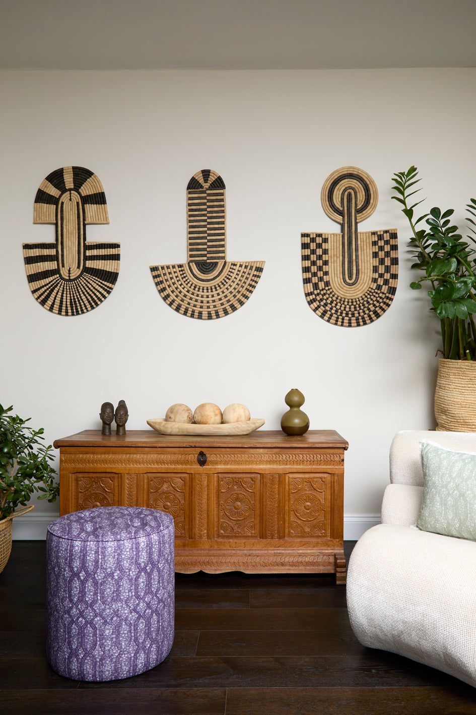 Living room scene with three black and tan sculptural woven panels, ornately carved wooden chest, baskets with house plans, white upholstered side chair and a stripe patterned footstool in purple.