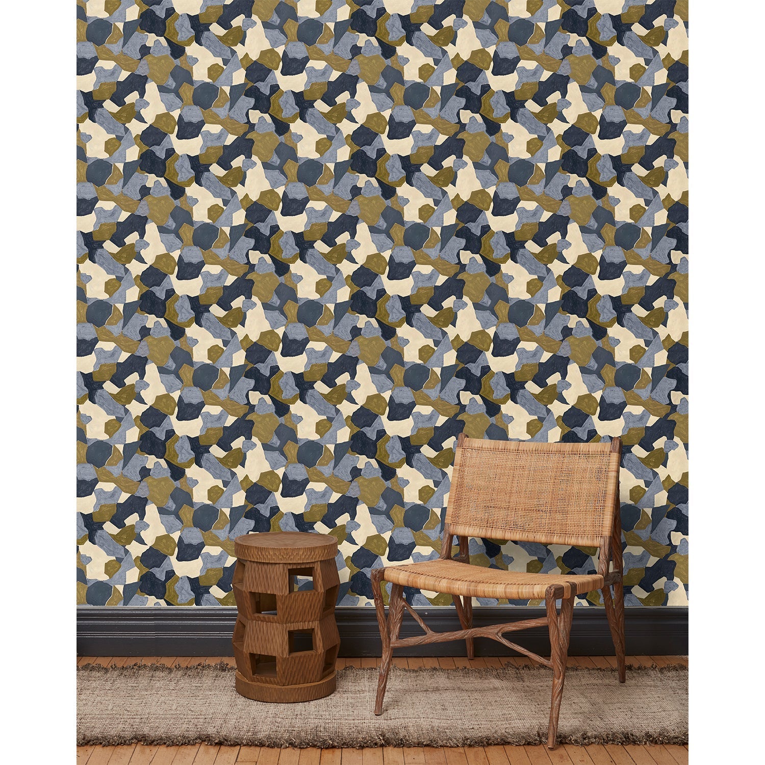 A wooden chair and end table in front of a wall papered in a painterly geometric pattern in shades of blue, cream and olive.