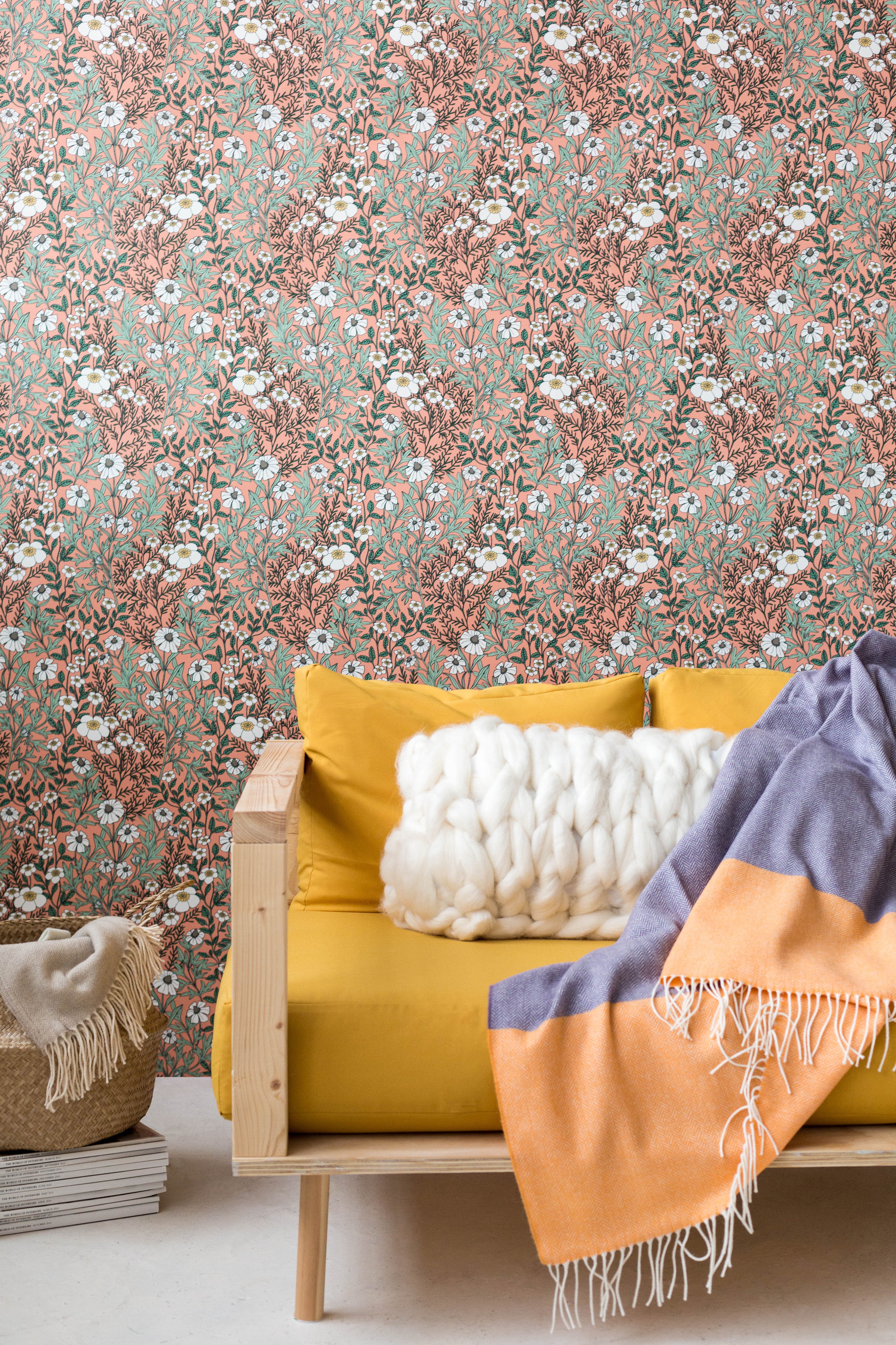 A couch covered in a throw pillow and blanket stands in front of a wall papered in a dense floral print in green and pink.