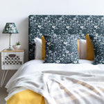 A bed with a headboard and pillows upholstered in a dense floral print in white, tan and green on a black field.