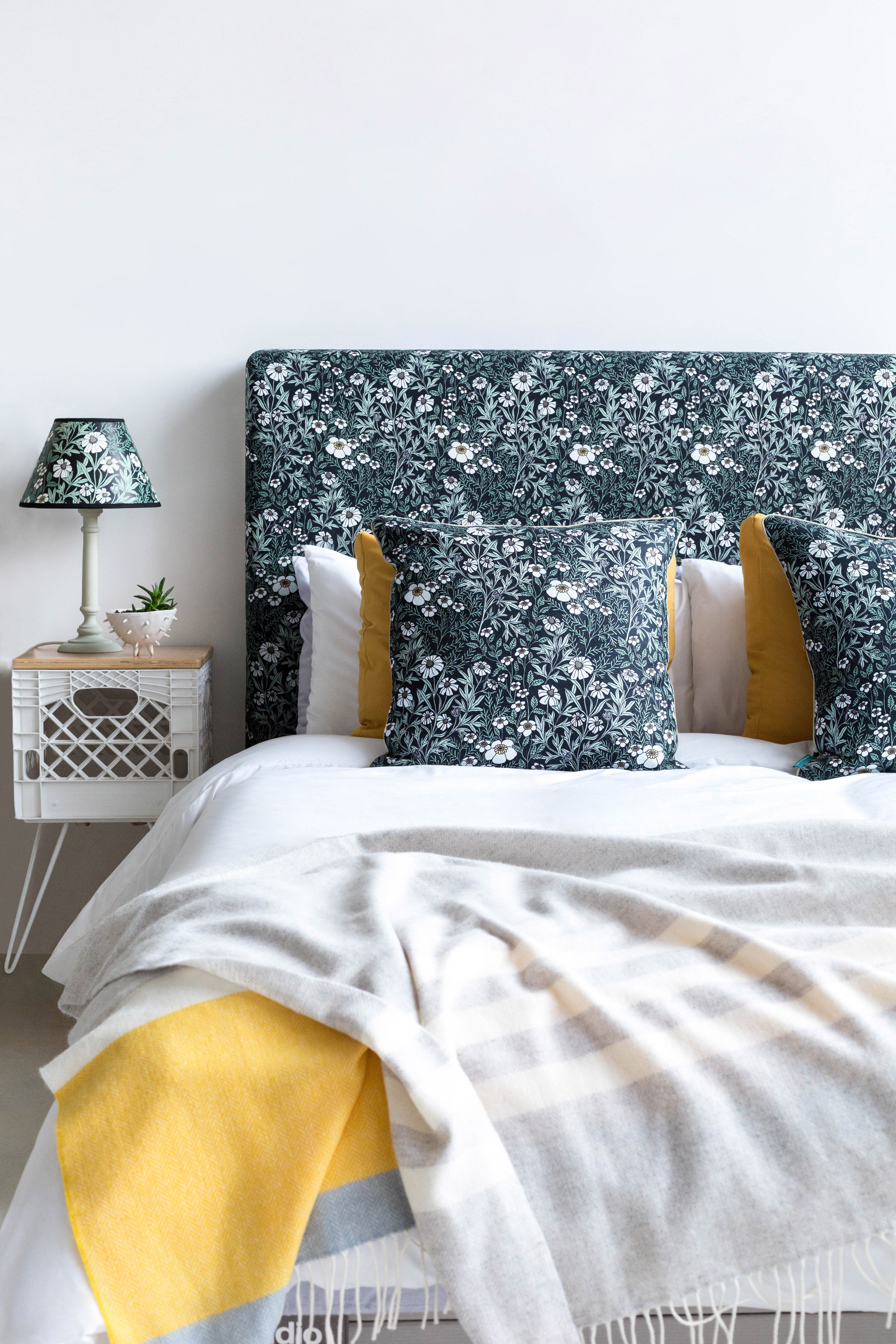 A bed with a headboard and pillows upholstered in a dense floral print in white, tan and green on a black field.