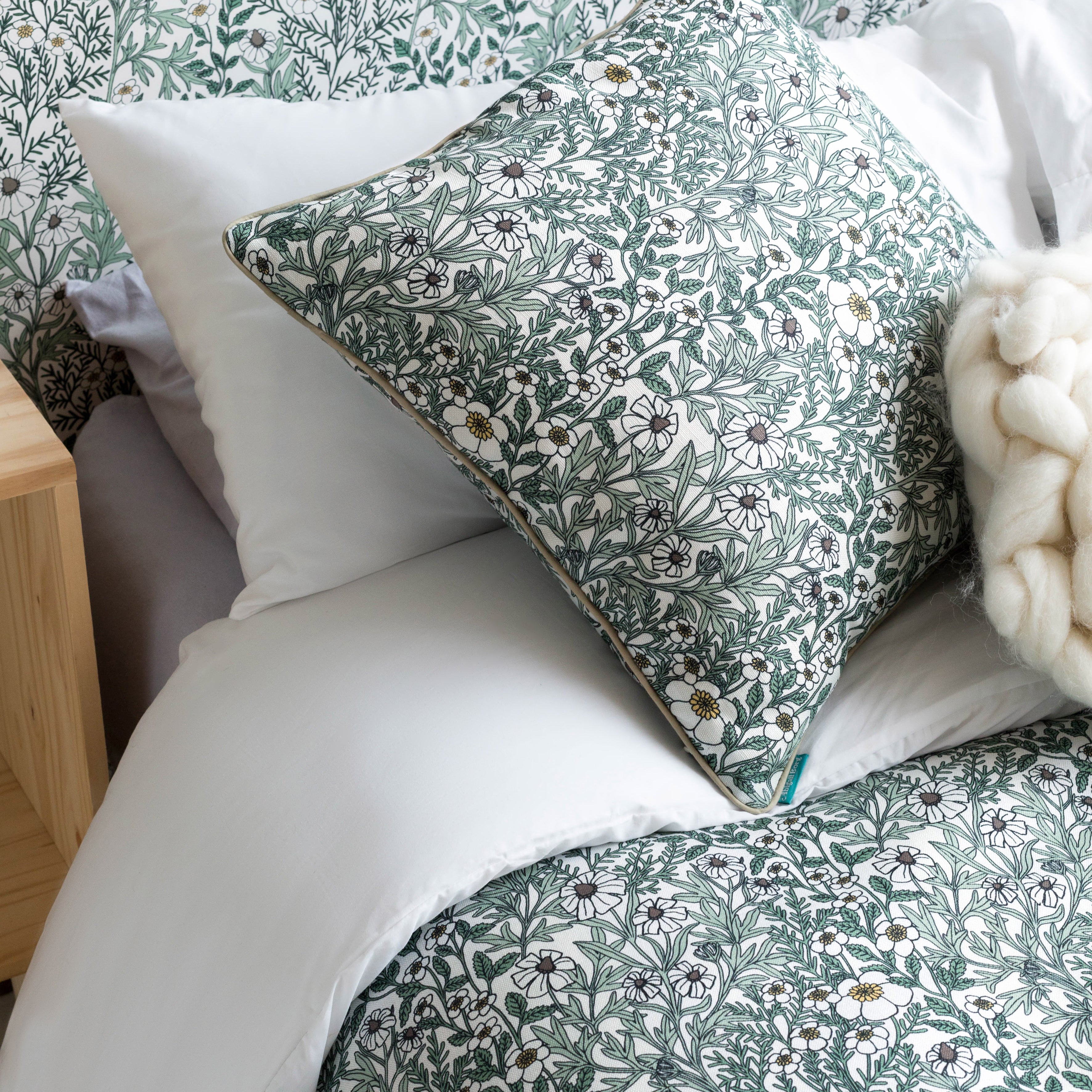 A bed with a headboard, duvet and pillows upholstered in a dense floral print in white, tan and green on a white field.