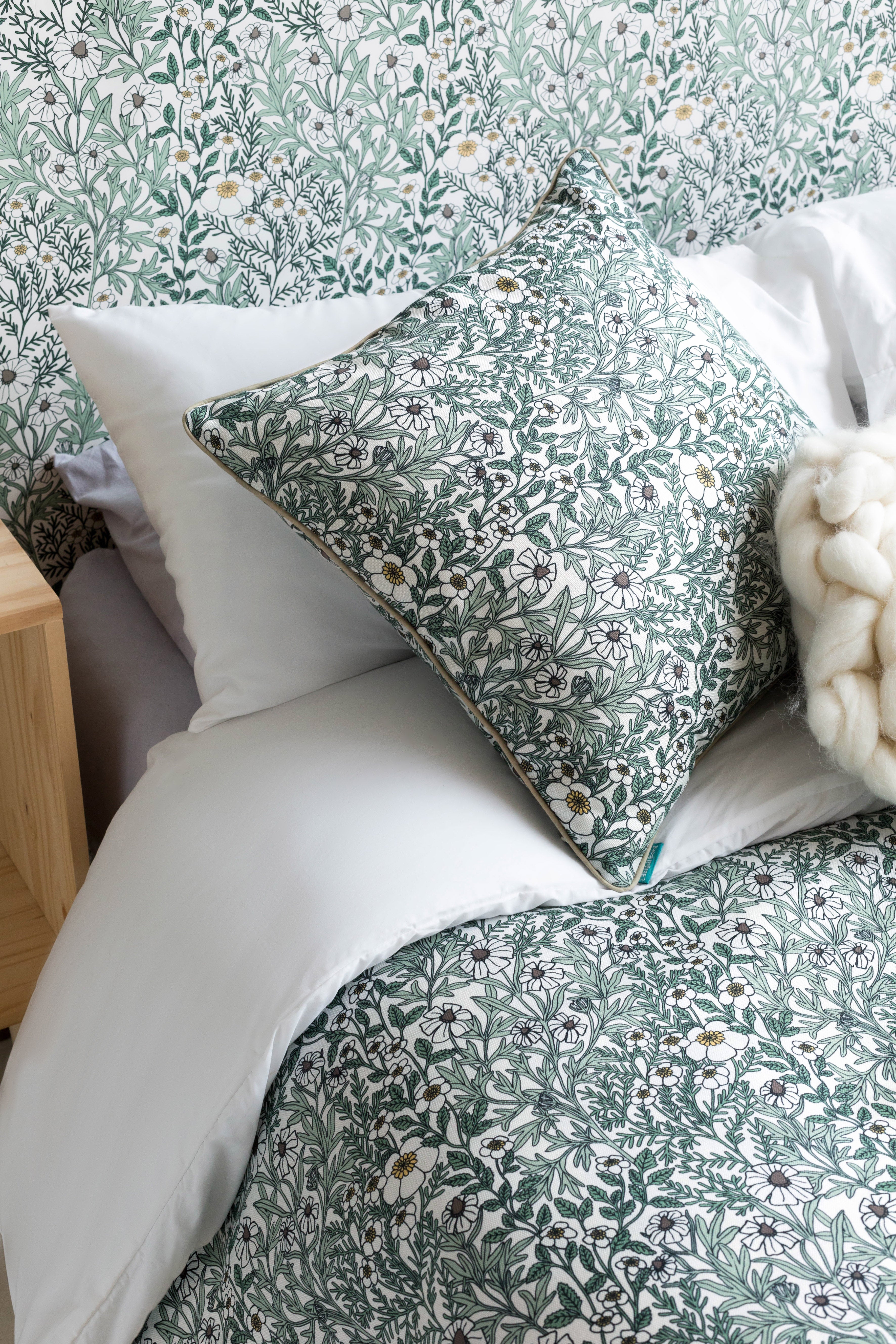 A bed with a headboard, duvet and pillows upholstered in a dense floral print in white, tan and green on a white field.