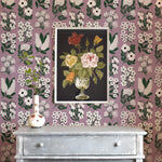 A dresser and flowers stands in front of a wall papered in a minimalist floral grid print in white, green and purple.