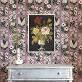 A dresser and flowers stands in front of a wall papered in a minimalist floral grid print in white, green and purple.