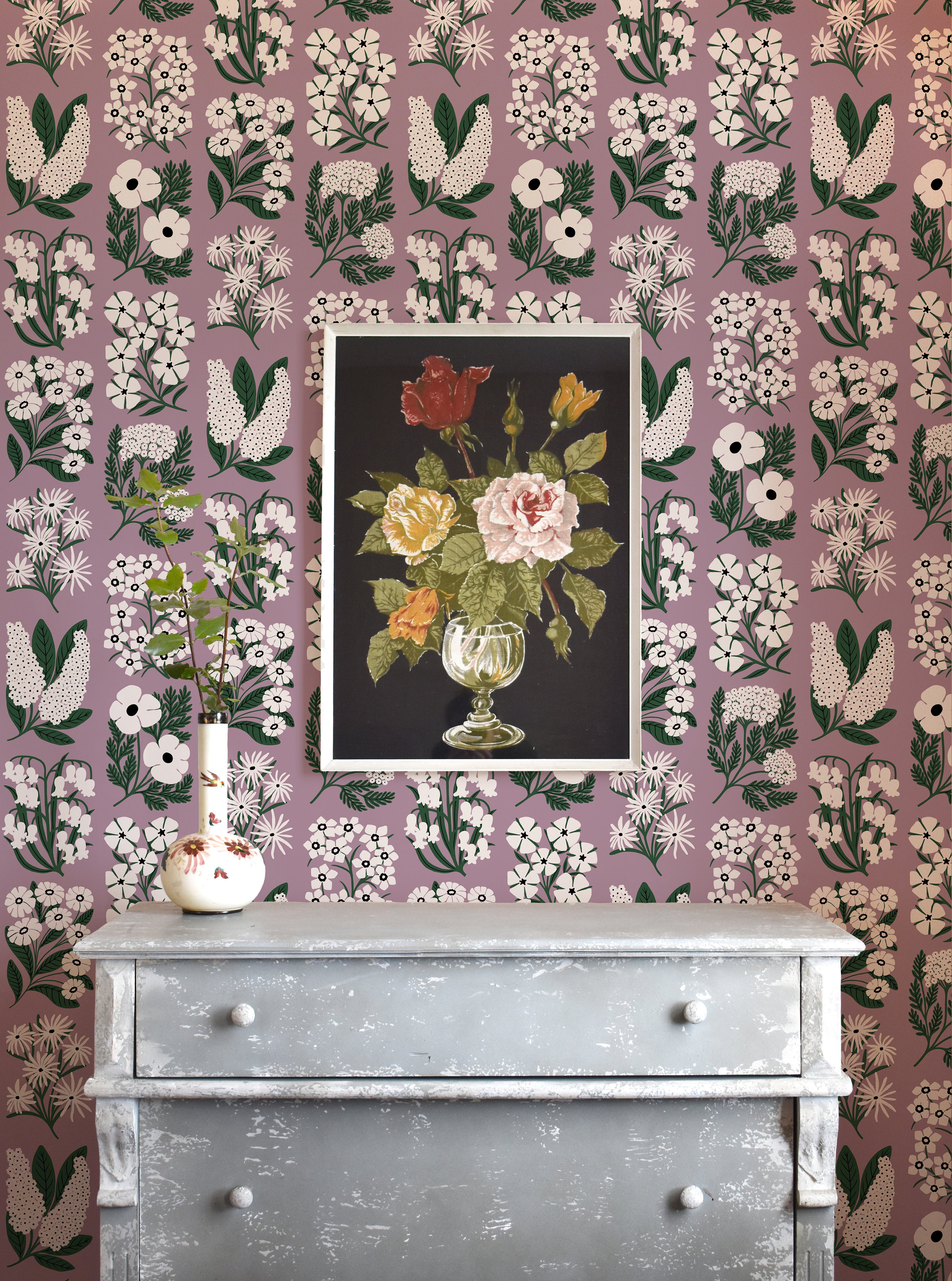A dresser and flowers stands in front of a wall papered in a minimalist floral grid print in white, green and purple.