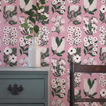 An end table and wooden chair stand in front of a wall papered in a minimalist floral grid print in white, green and pink.