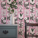 An end table and wooden chair stand in front of a wall papered in a minimalist floral grid print in white, green and pink.