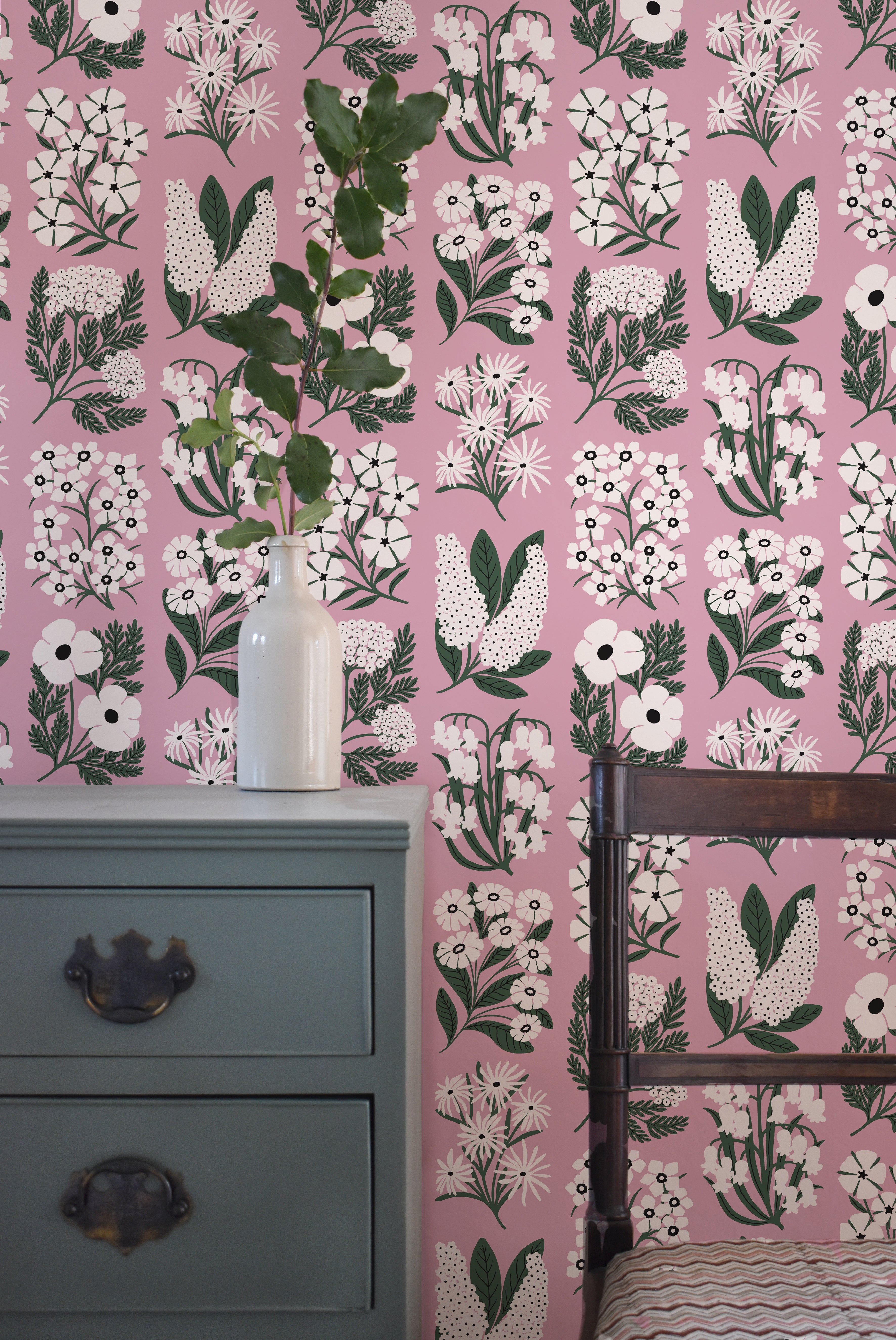 An end table and wooden chair stand in front of a wall papered in a minimalist floral grid print in white, green and pink.