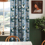 A cozy dining room with a large window and curtains in a minimalist floral grid print in white and green on a blue field.