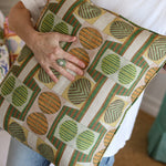Close up of a woman in white tee shirt and jeans holding a bold geometric patterned pillow in orange, brown and green. 