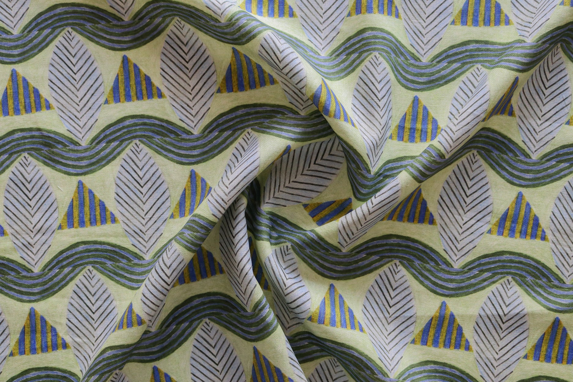 A close-up of a gently draped patterned fabric featuring repeating rows of stylized grey leaves, blue and yellow stripe triangles and wavy blue-yellow lines on yellow background.