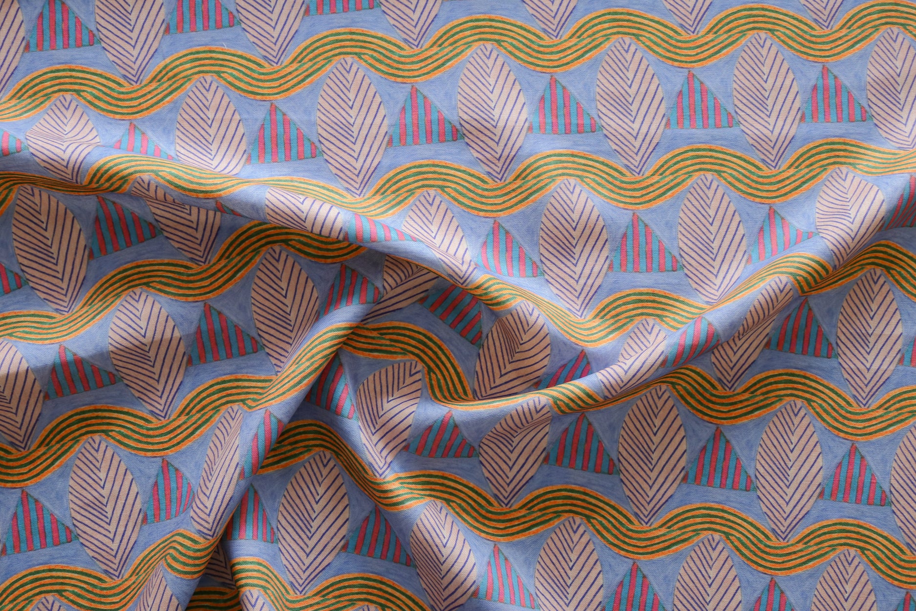 A close-up of a gently draped patterned fabric featuring repeating rows of stylized pink leaves, blue and red striped triangles and wavy orange-green lines on a blue background