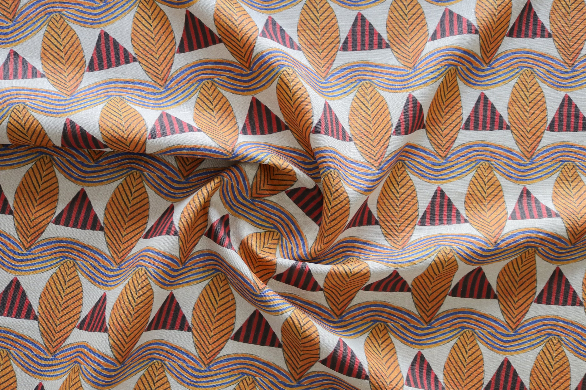 A close-up of a gently draped patterned fabric featuring repeating rows of stylized orange leaves, navy and red striped triangles and wavy orange-blue lines on a white background