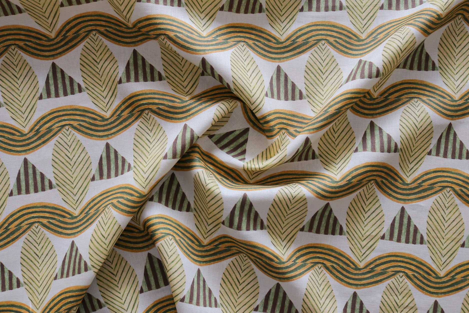 A close-up of a gently draped patterned fabric featuring repeating rows of stylized tan leaves, brown and green striped triangles and wavy tan-green lines on a white background