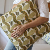 Detail of a woman in a white shirt and jeans holding a bold abstract leaf patterned throw pillow in tan and green in front of a white couch. 