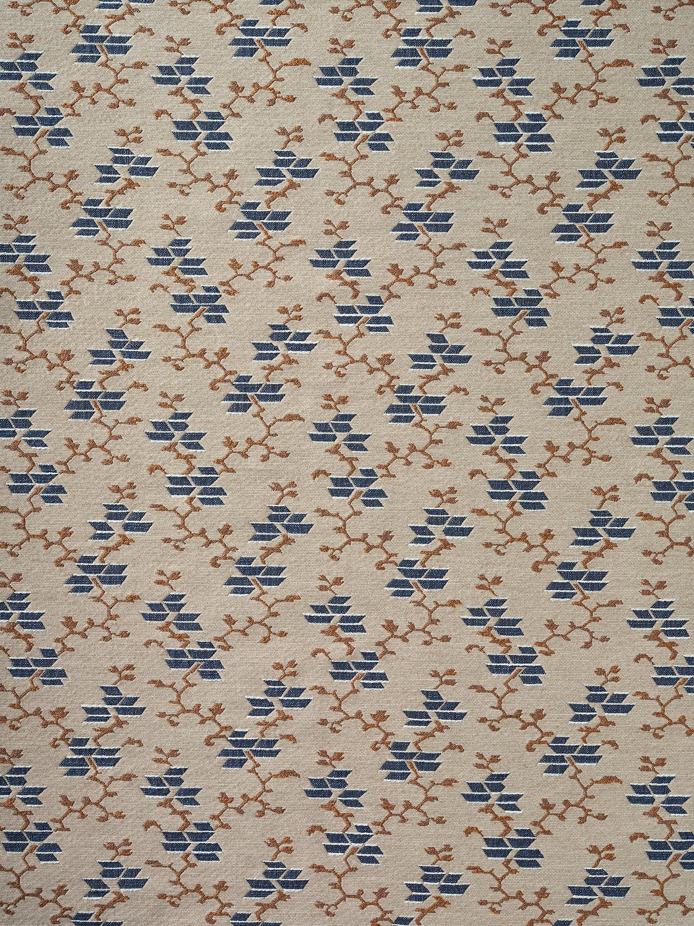 A close-up of a woven fabric pattern featuring a repeating geometric design of blue diamond floral shapes and brown branch motifs on a warm beige background.