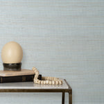 End table with books and a lamp in front of a wall papered in textured grasscloth in shades of pastel, blue and turquoise.