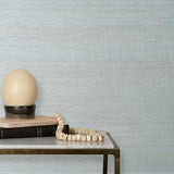 End table with books and a lamp in front of a wall papered in textured grasscloth in shades of pastel, blue and turquoise.