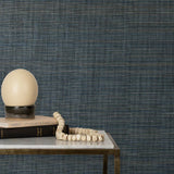 End table with books and a lamp in front of a wall papered in textured grasscloth in shades of green, cream and turquoise.