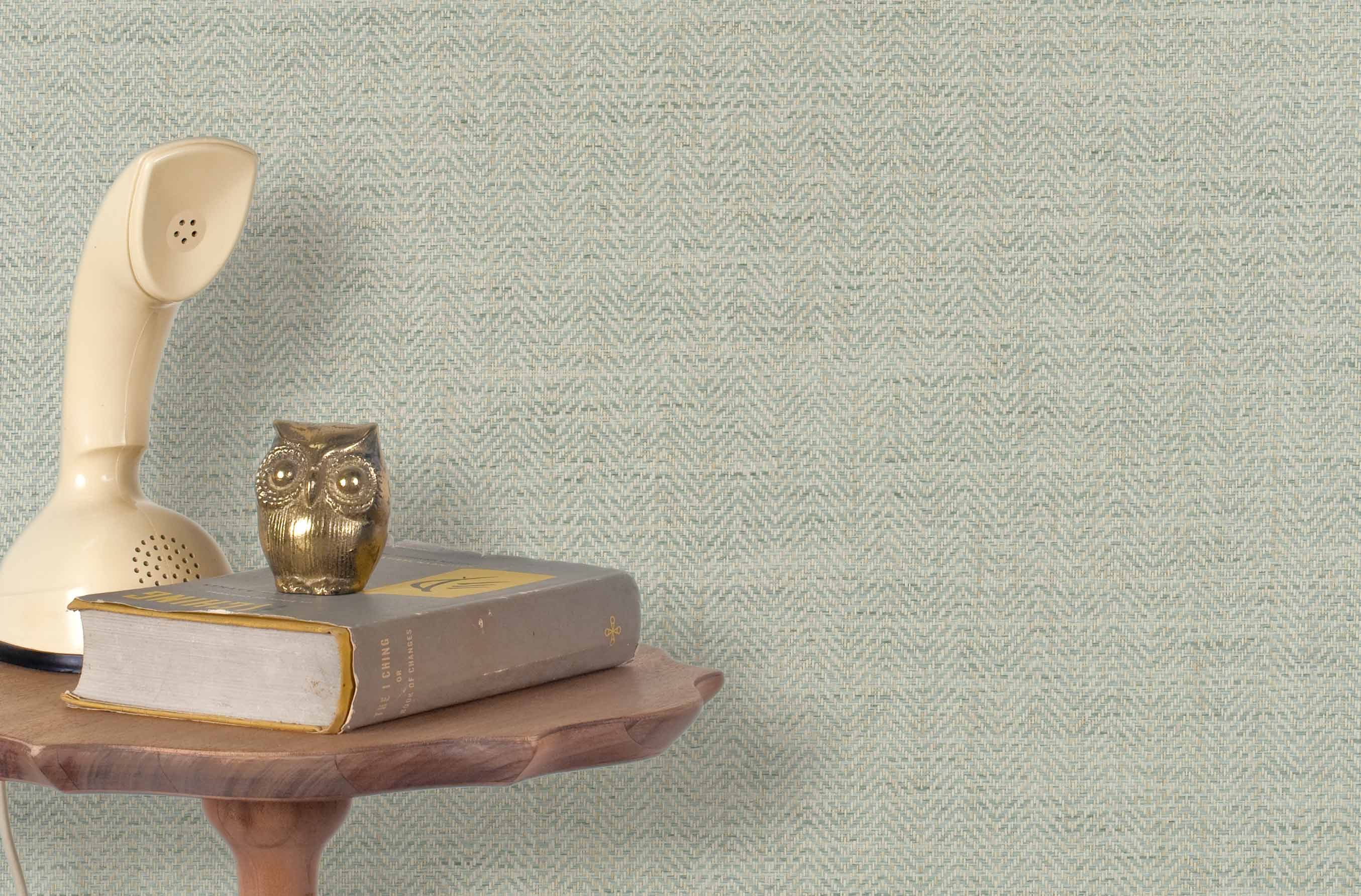 End table with books and  vintage phone in front of a grasscloth wallpaper with a herringbone weave in cream and aqua.
