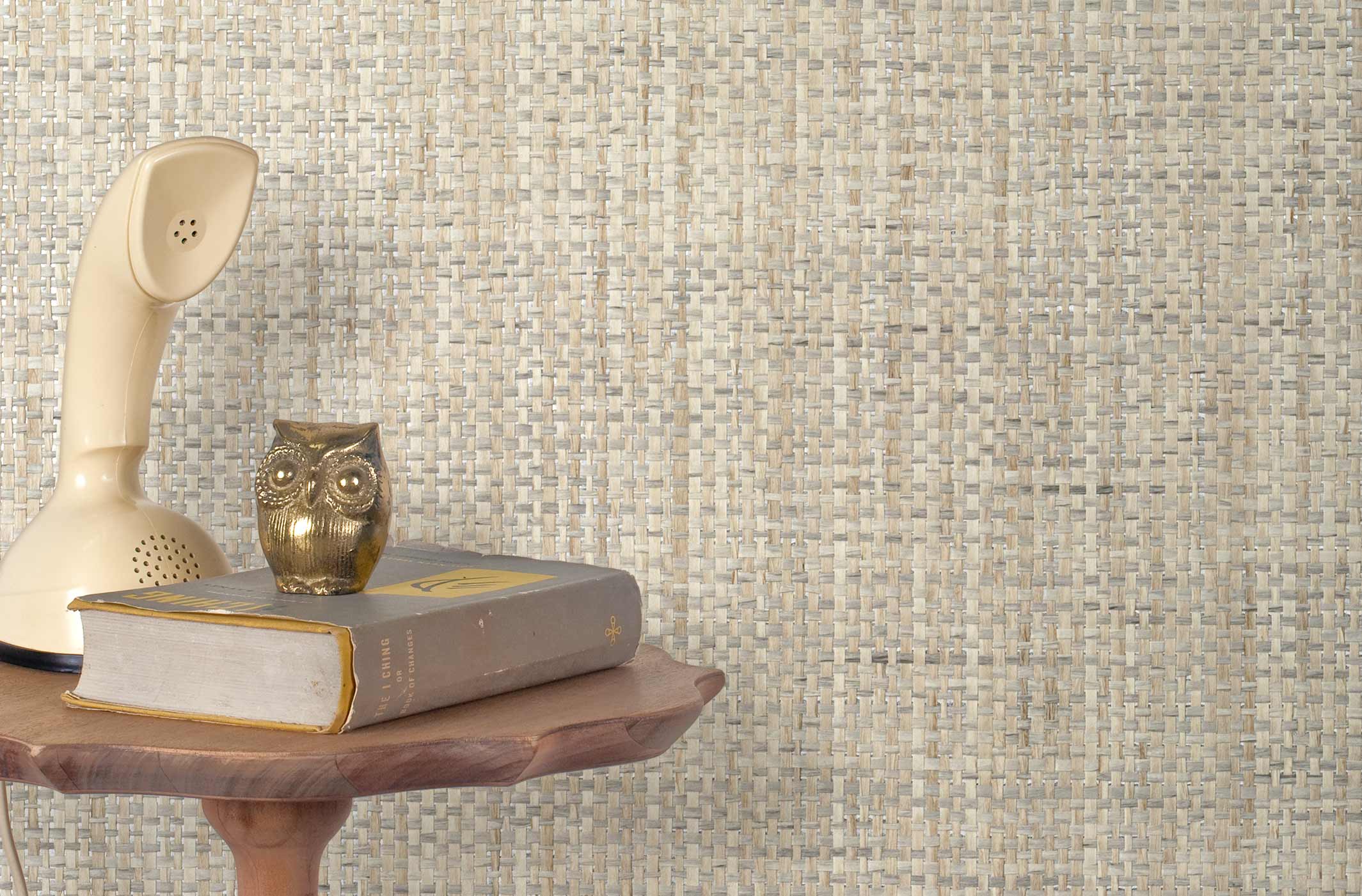 End table with a phone, book and owl sculpture in front of a wall covered in paperweave grasscloth in mottled cream and gray.