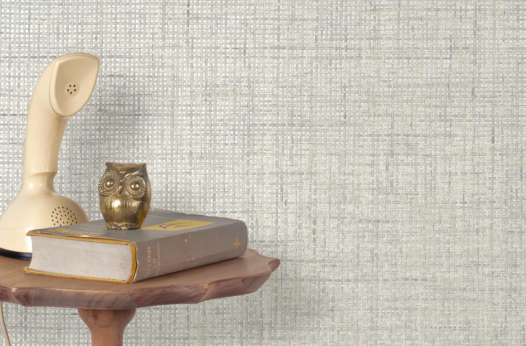 End table with a phone, book and owl sculpture in front of a wall covered in paperweave grasscloth in metallic cream and gray.