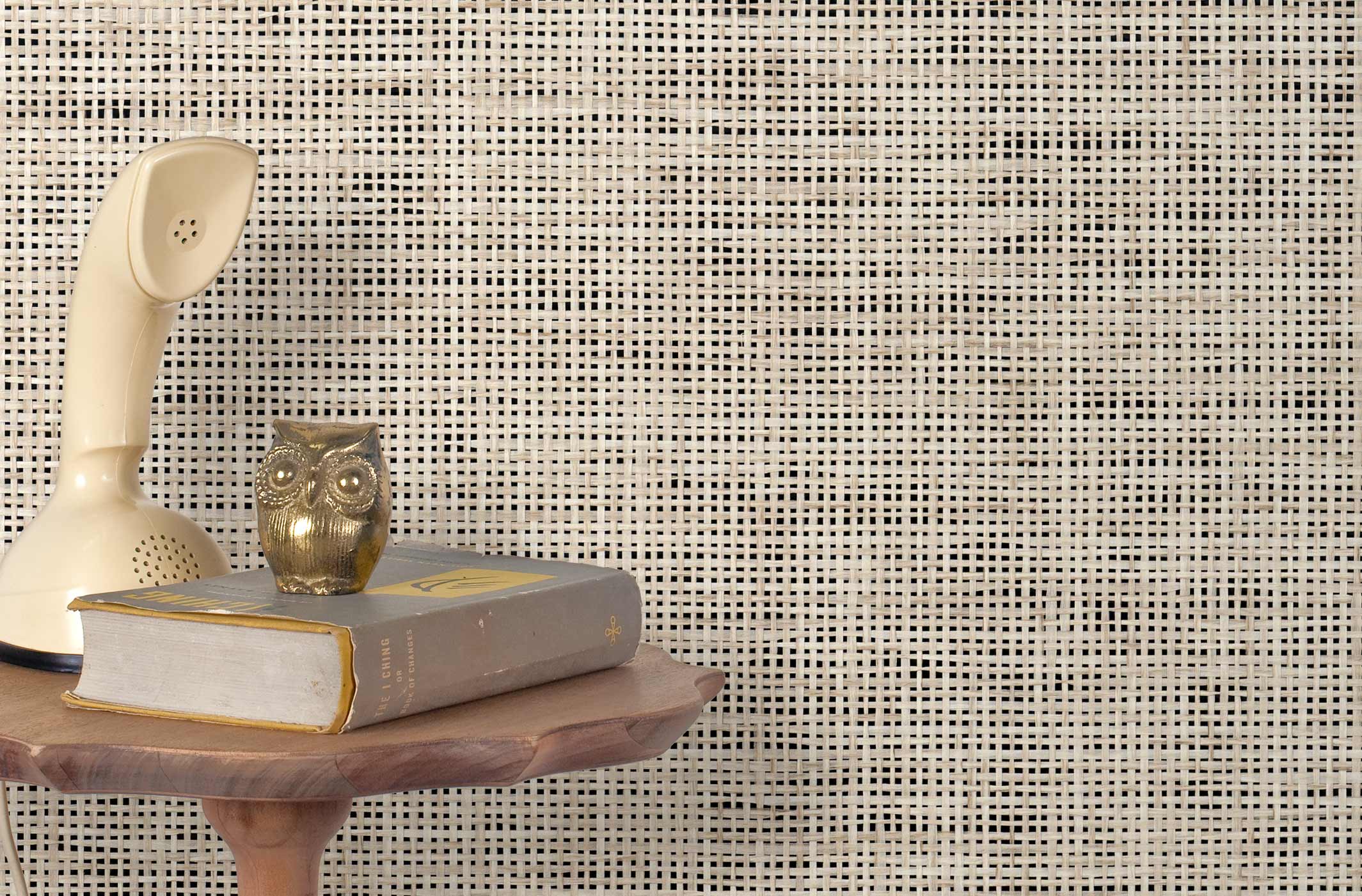 End table with a phone, book and owl sculpture in front of a wall covered in paperweave grasscloth in mottled cream and black.
