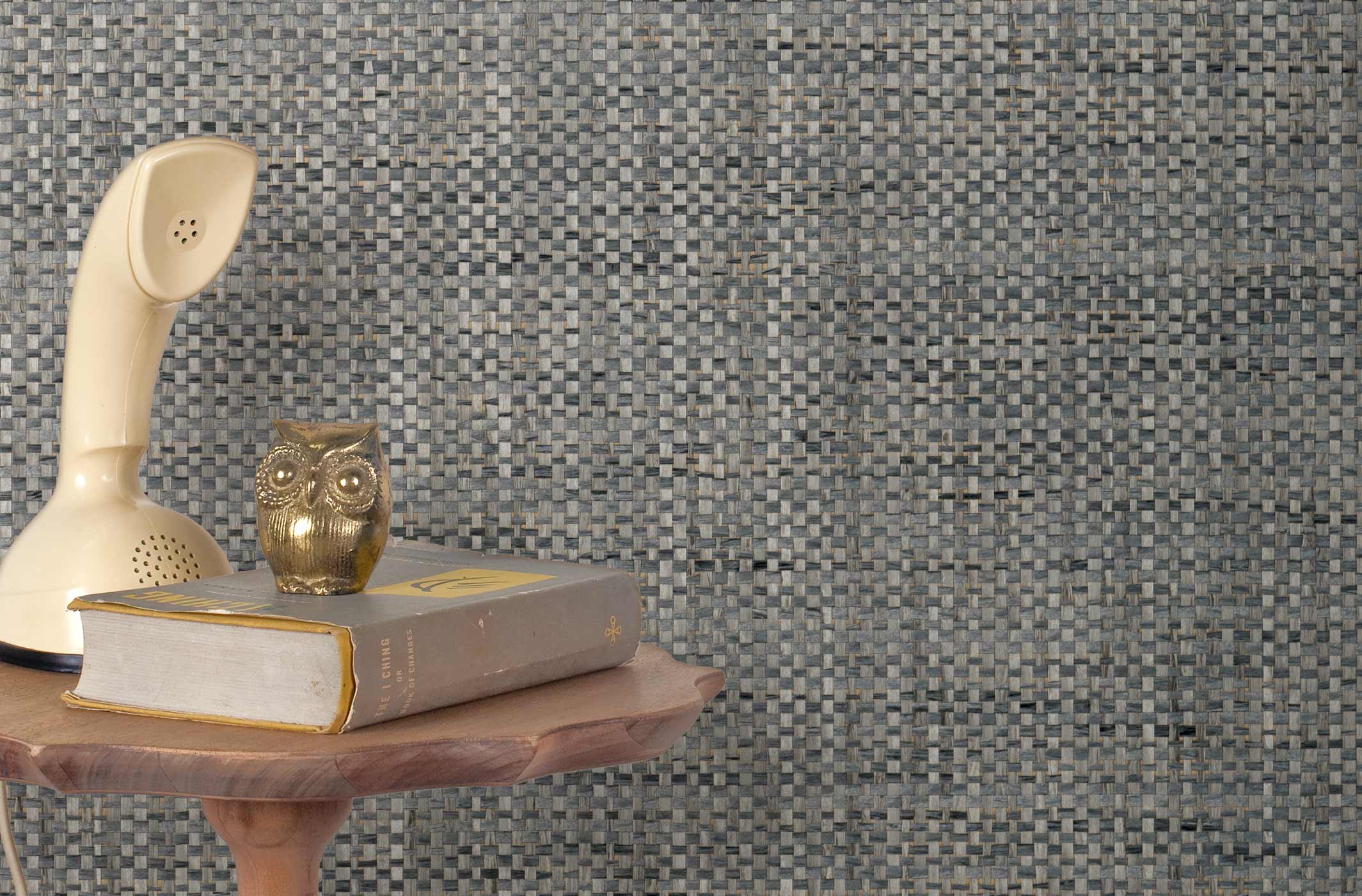 End table with a phone, book and owl sculpture in front of a wall covered in paperweave grasscloth in mottled blue-gray and tan.