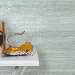 End table with a vase and sculptures in front of a wall papered in metallic-rubbed jute grasscloth in blue pastel.