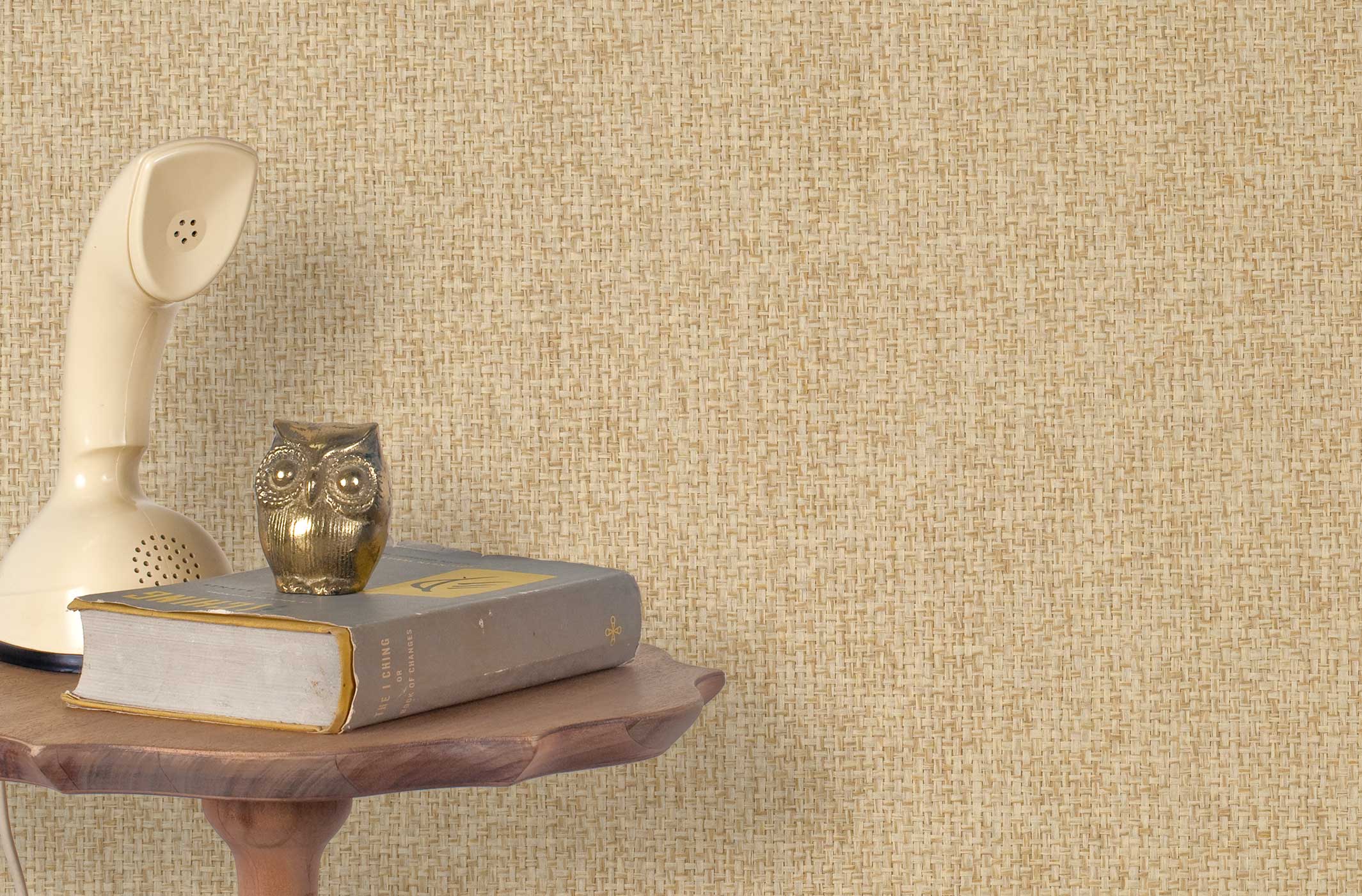 End table with a phone, book and owl sculpture in front of a wall covered in paperweave grasscloth in mottled tan.