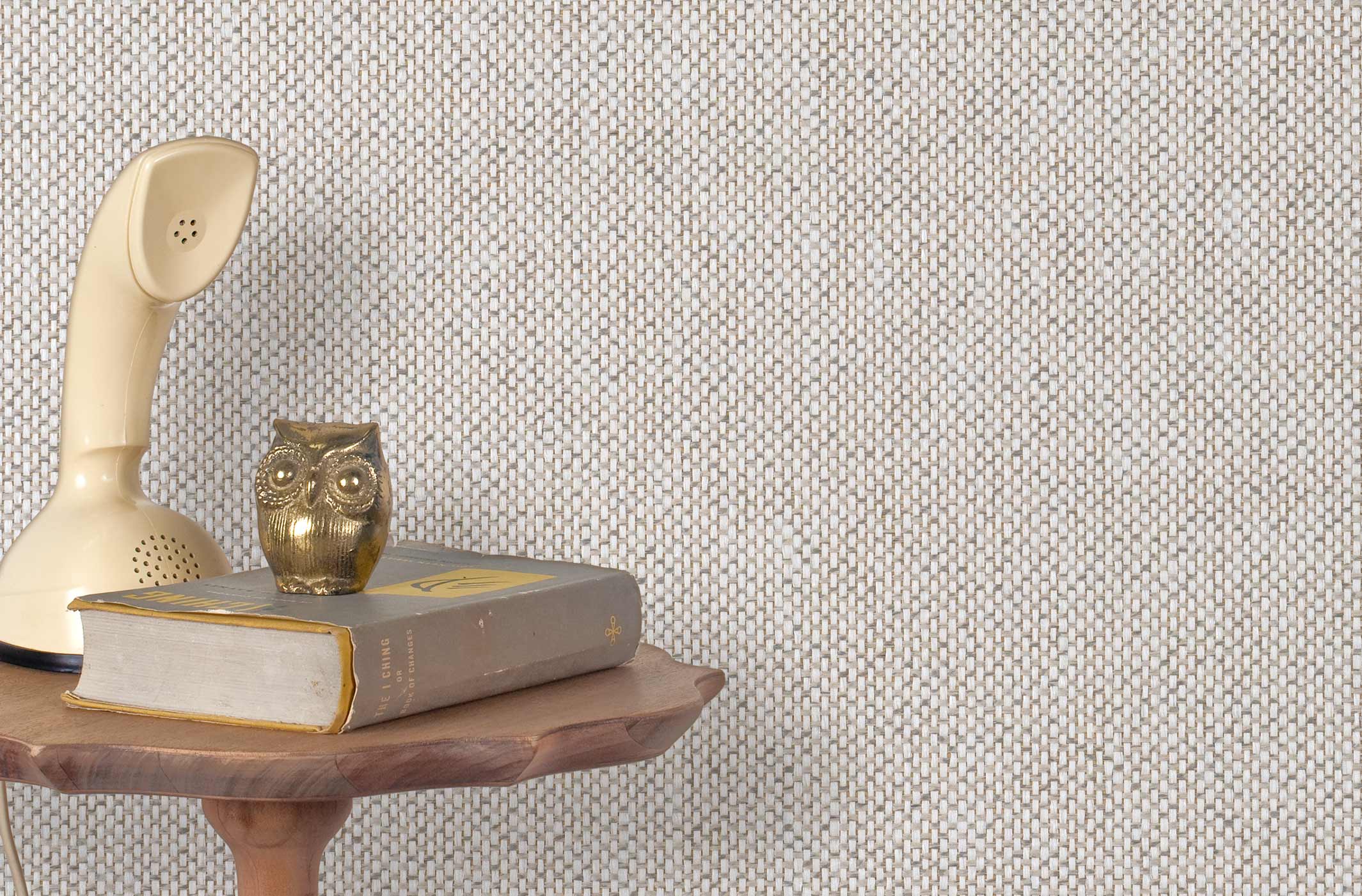 End table with a phone, book and owl sculpture in front of a wall covered in paperweave grasscloth in checked cream and gray.