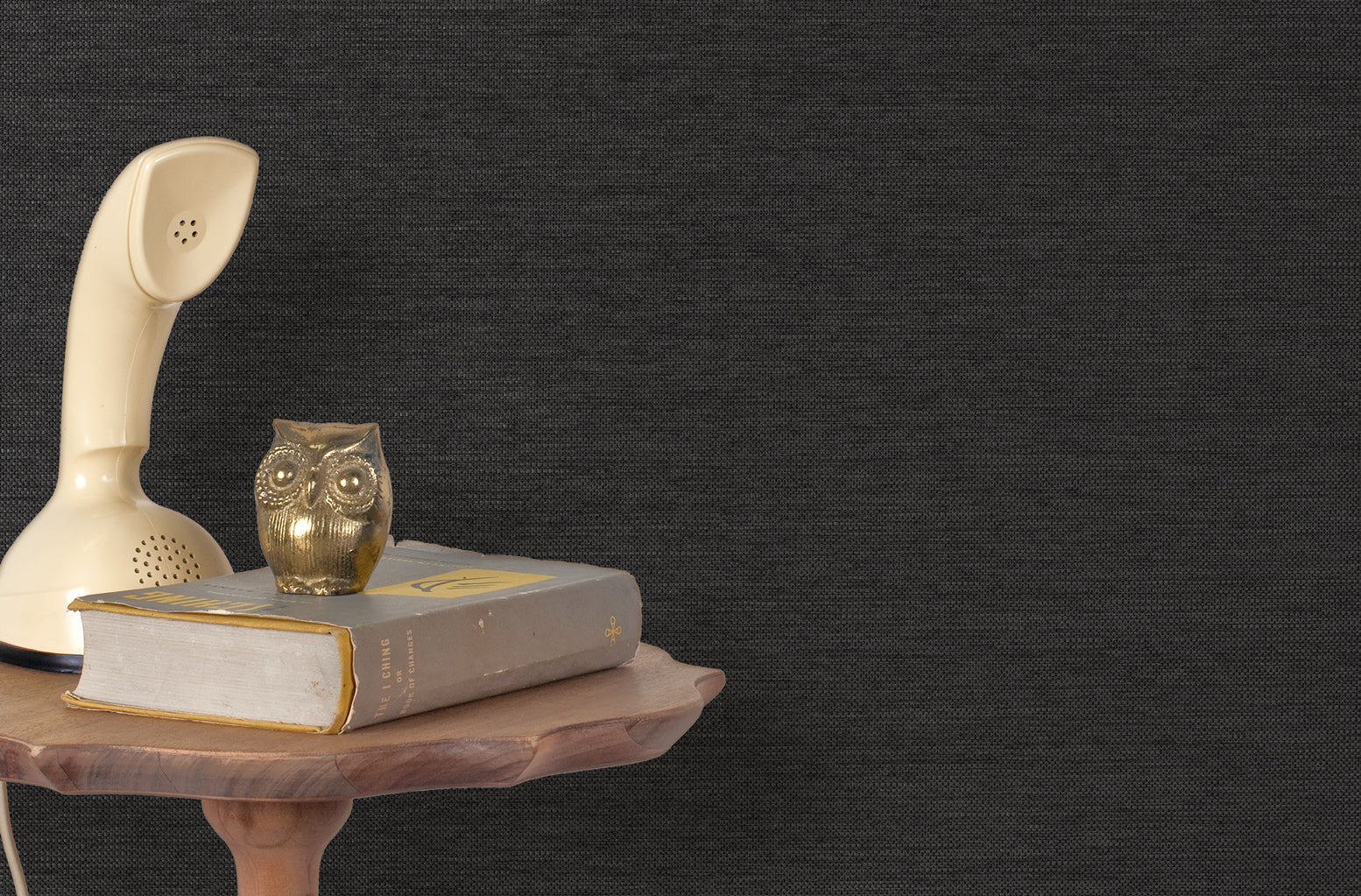 End table with a phone, book and owl sculpture in front of a wall covered in paperweave grasscloth in mottled charcoal.