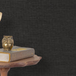 End table with a phone, book and owl sculpture in front of a wall covered in paperweave grasscloth in mottled charcoal.