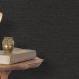 End table with a phone, book and owl sculpture in front of a wall covered in paperweave grasscloth in mottled charcoal.