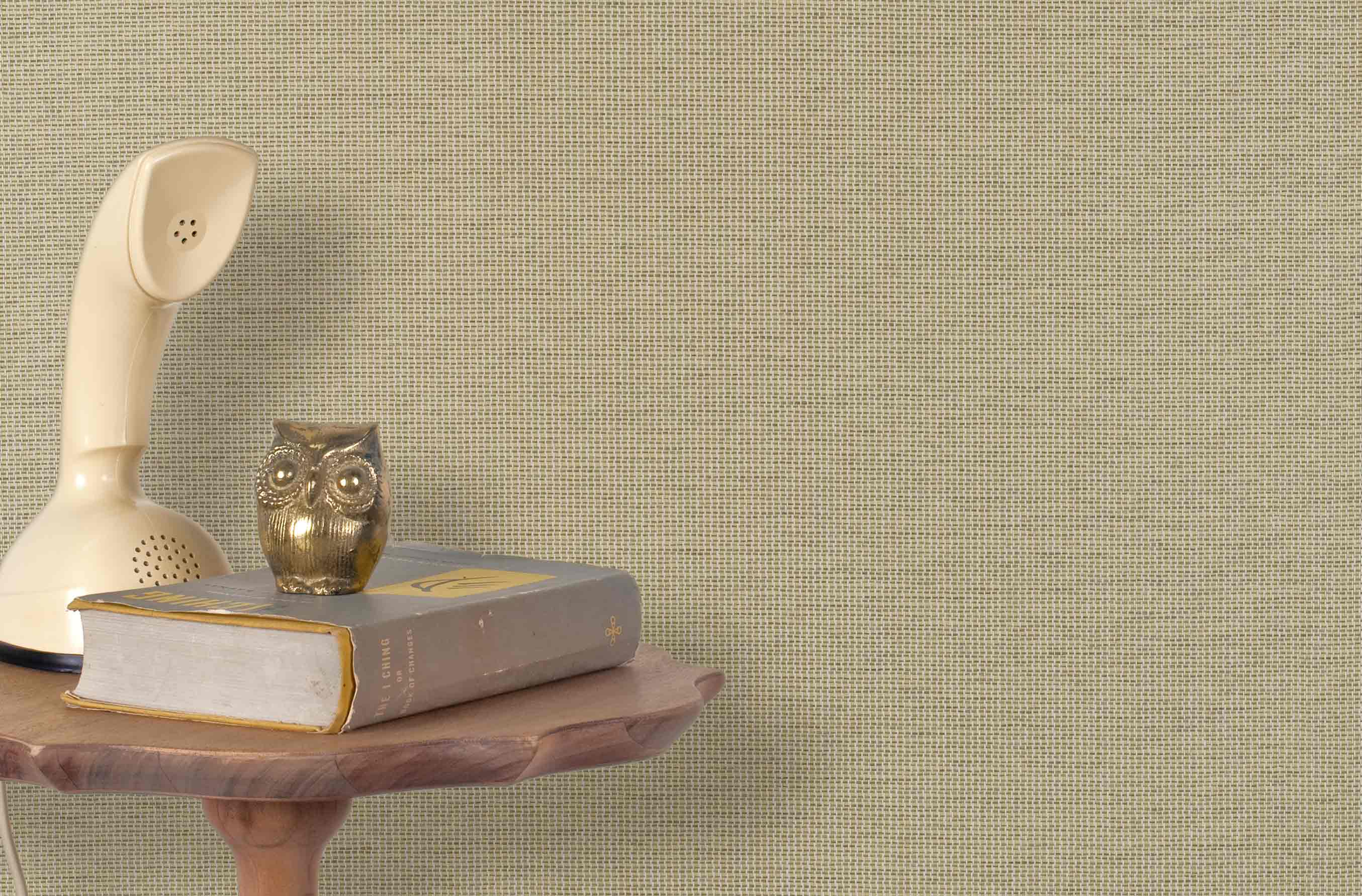 End table with a phone, book and owl sculpture in front of a wall covered in paperweave grasscloth in natural taupe.