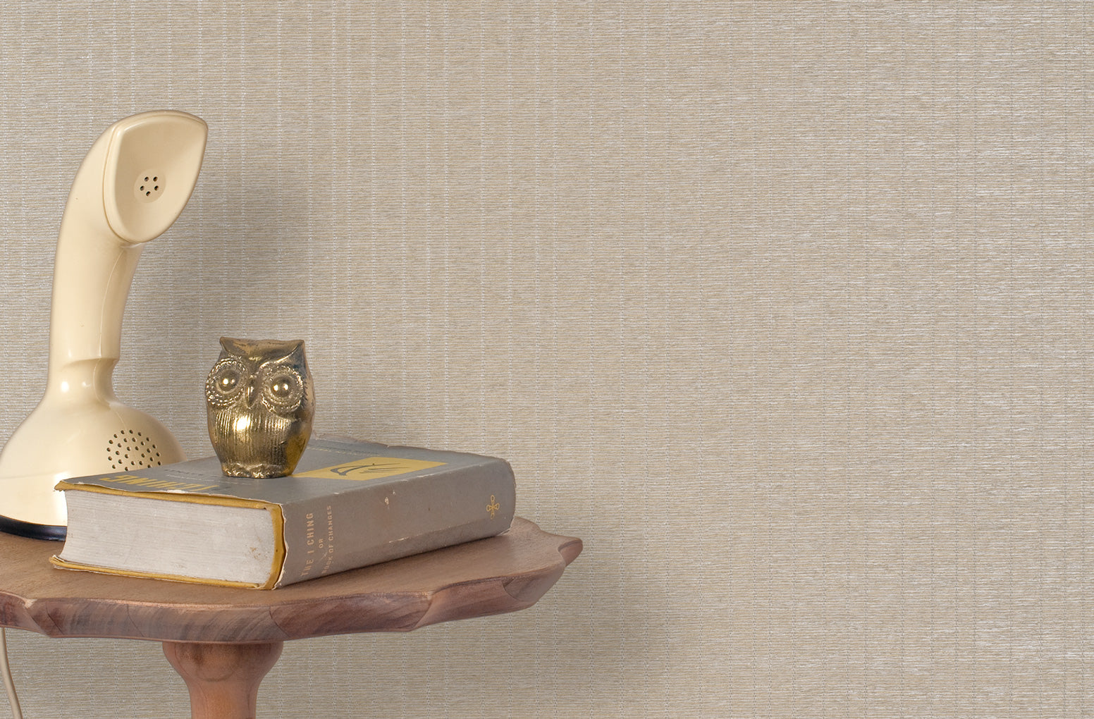 End table with a phone, book and owl sculpture in front of a wall covered in metallic paperweave grasscloth in tan.