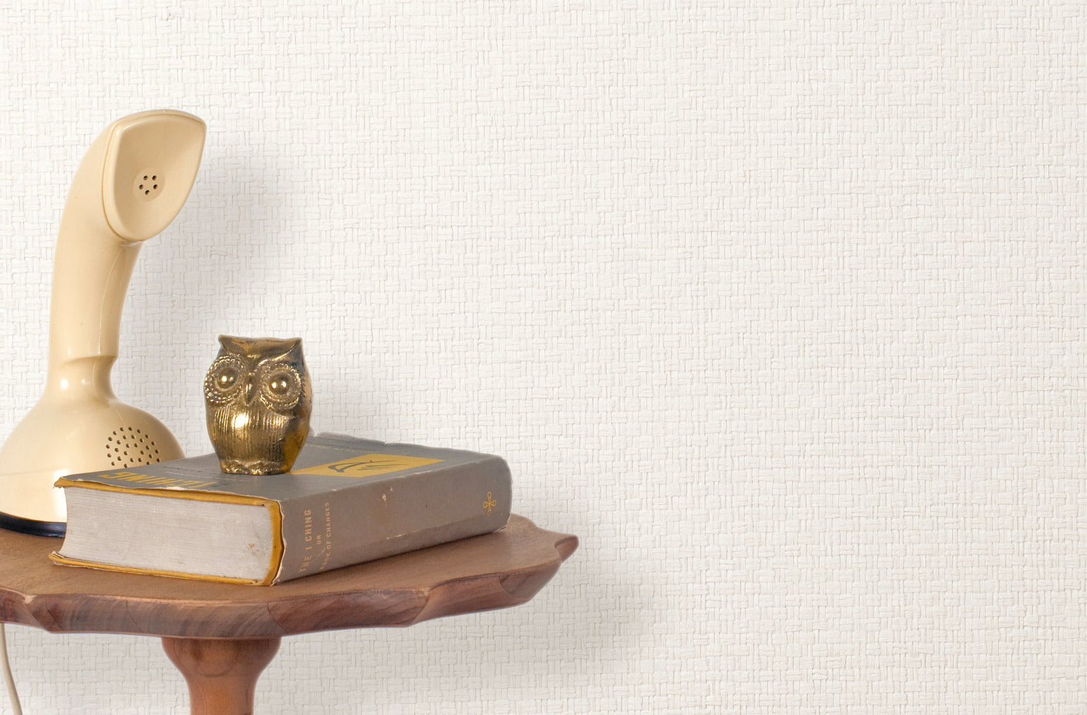 End table with a phone, book and owl sculpture in front of a wall covered in paperweave grasscloth in white.