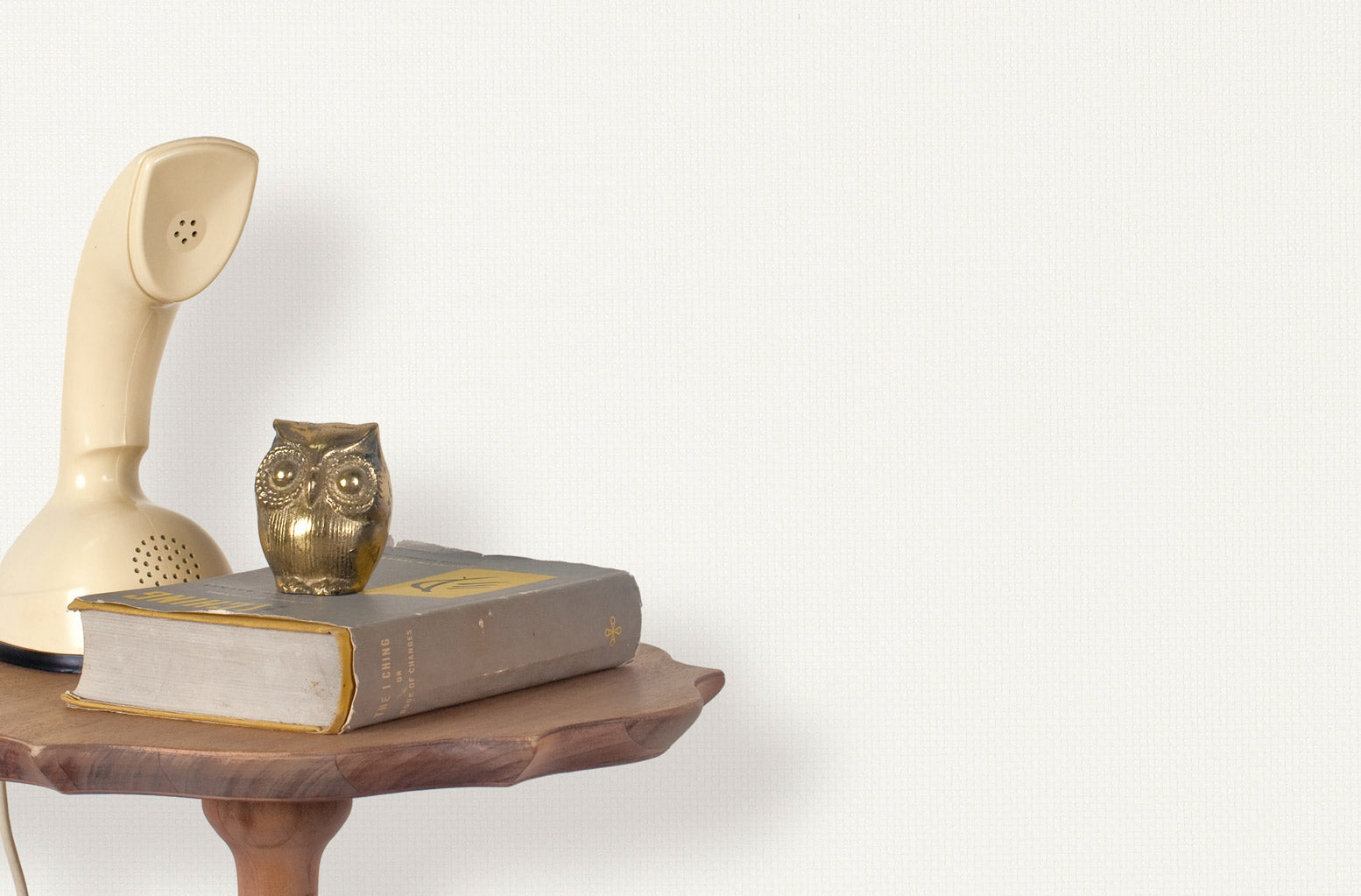 End table with a phone, book and owl sculpture in front of a wall covered in paperweave grasscloth in white.