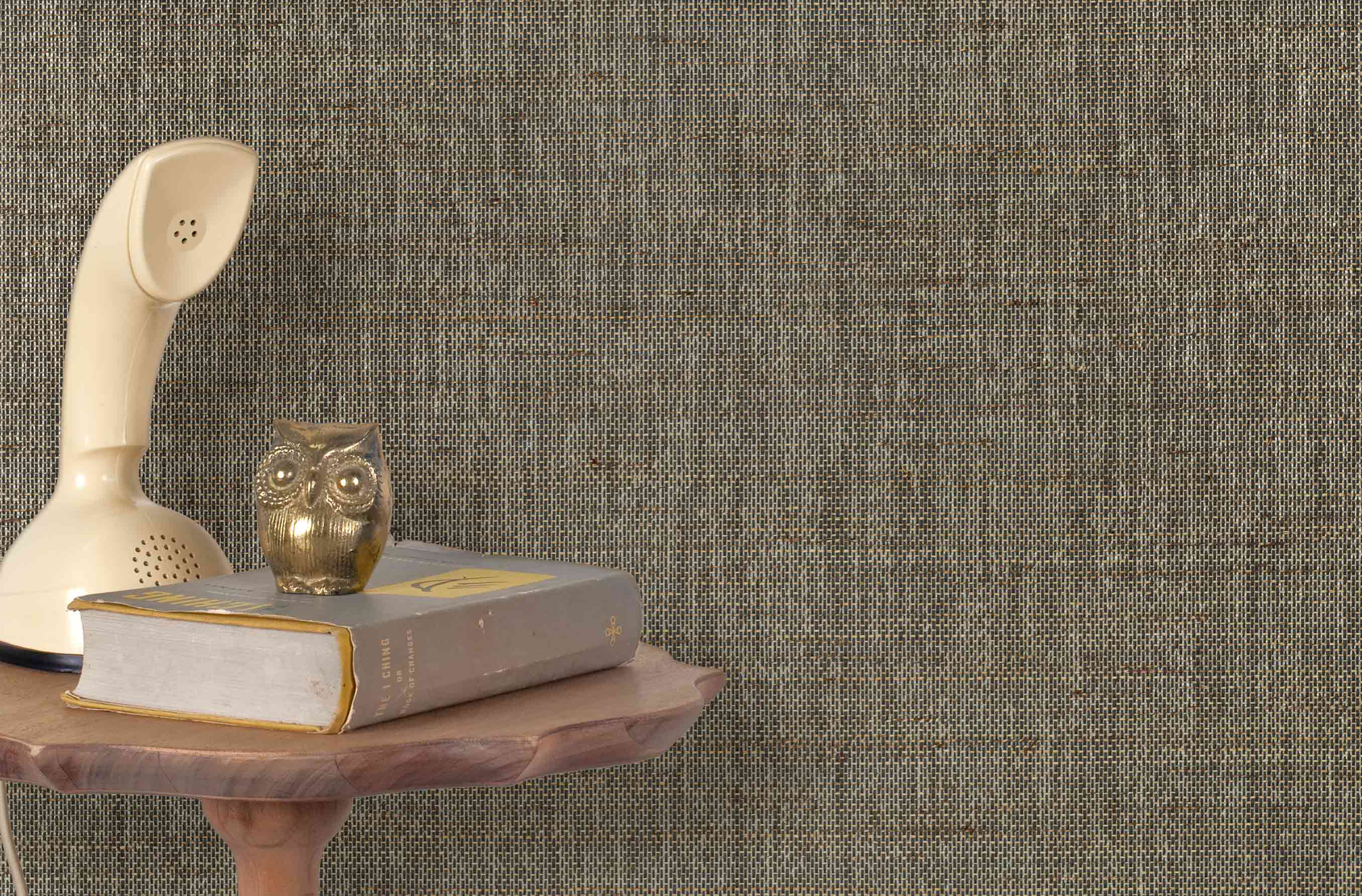 End table with a phone, book and owl sculpture in front of a wall covered in paperweave grasscloth in mottled brown.