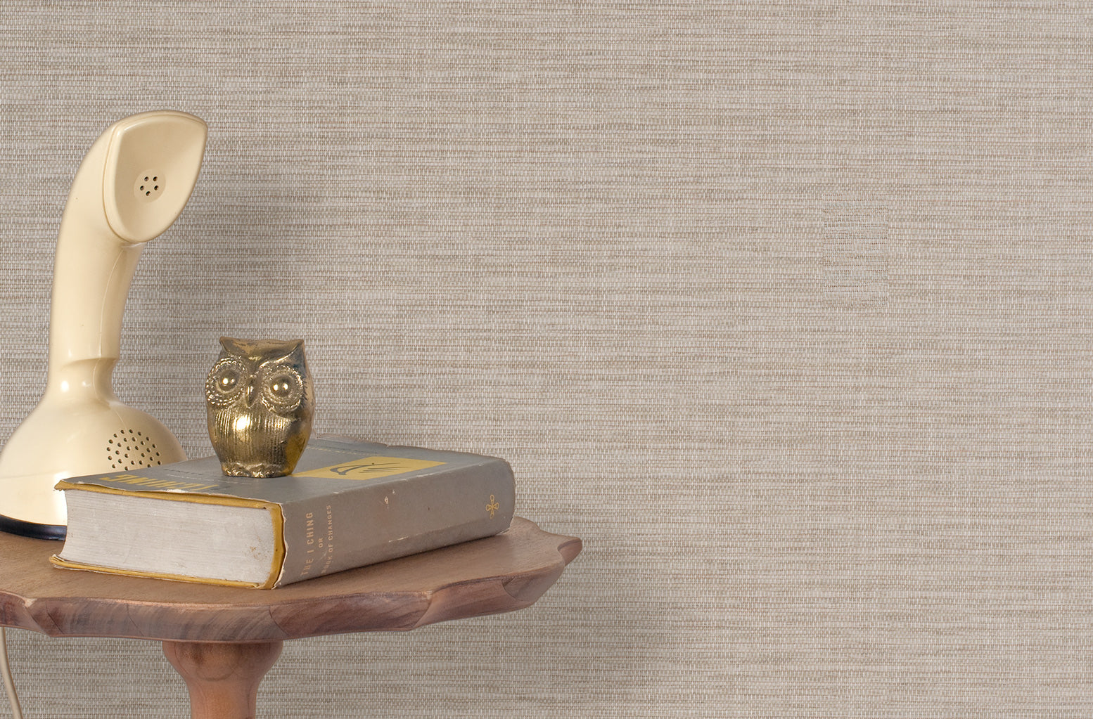 End table with a phone, book and owl sculpture in front of a wall covered in paperweave grasscloth in neutral.