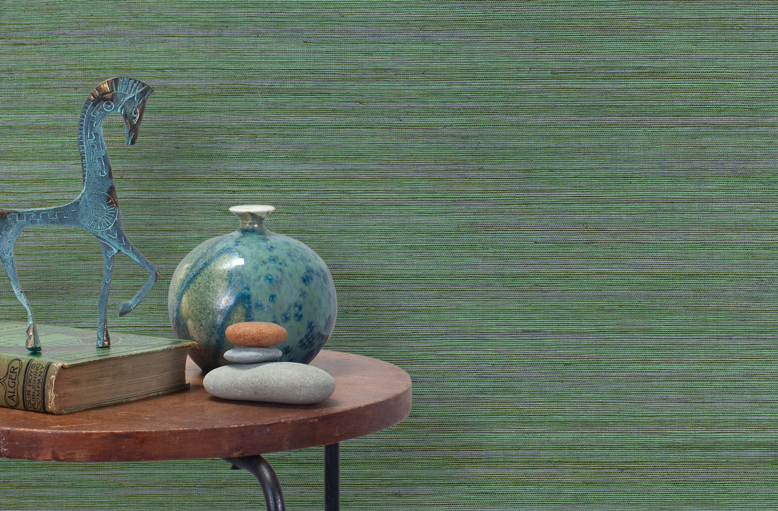 End table with a vase, horse sculpture and stones in front of a wall covered in sisal grasscloth in mottled green.