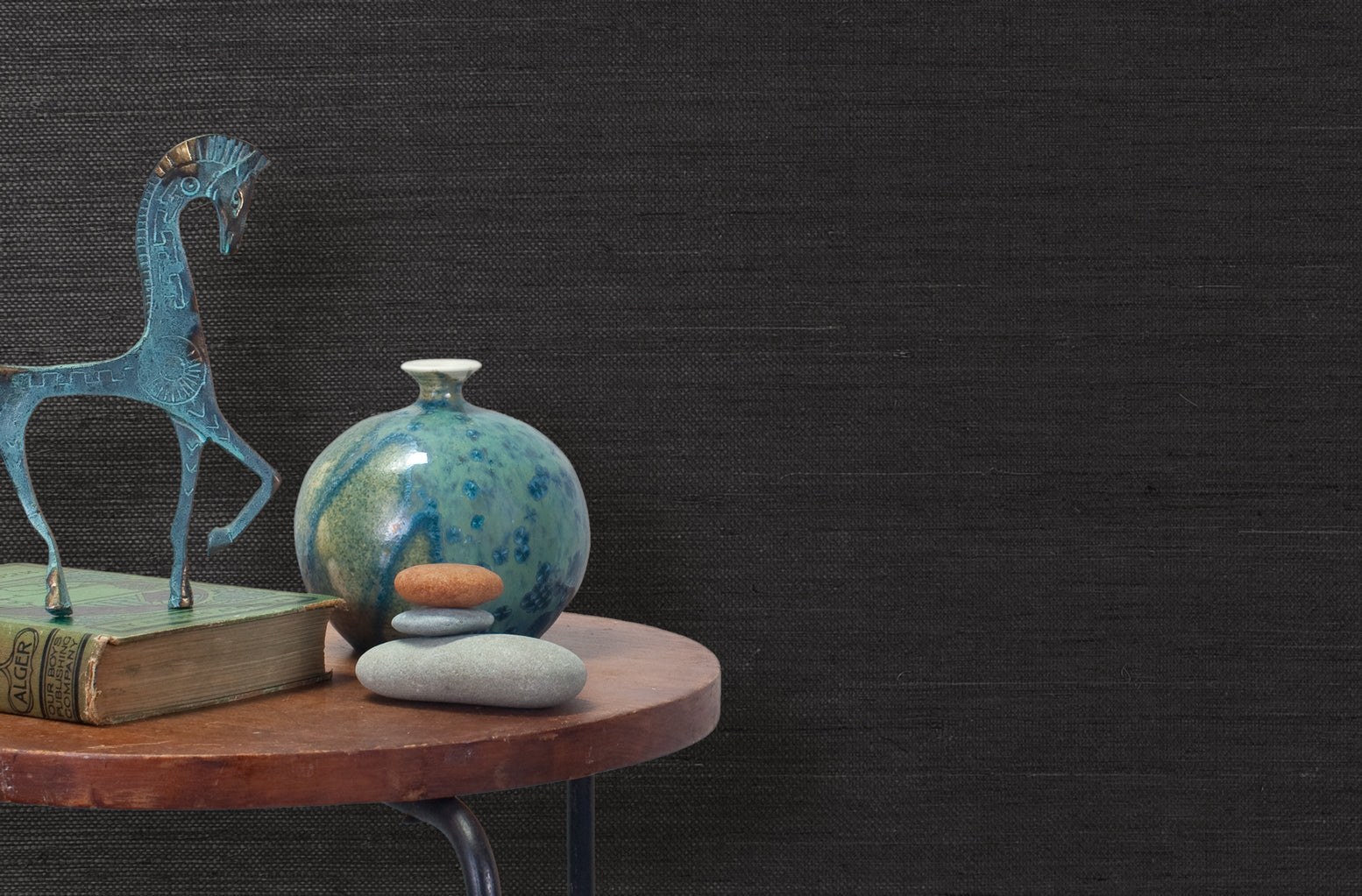 End table with a vase, horse sculpture and stones in front of a wall covered in sisal grasscloth in black.