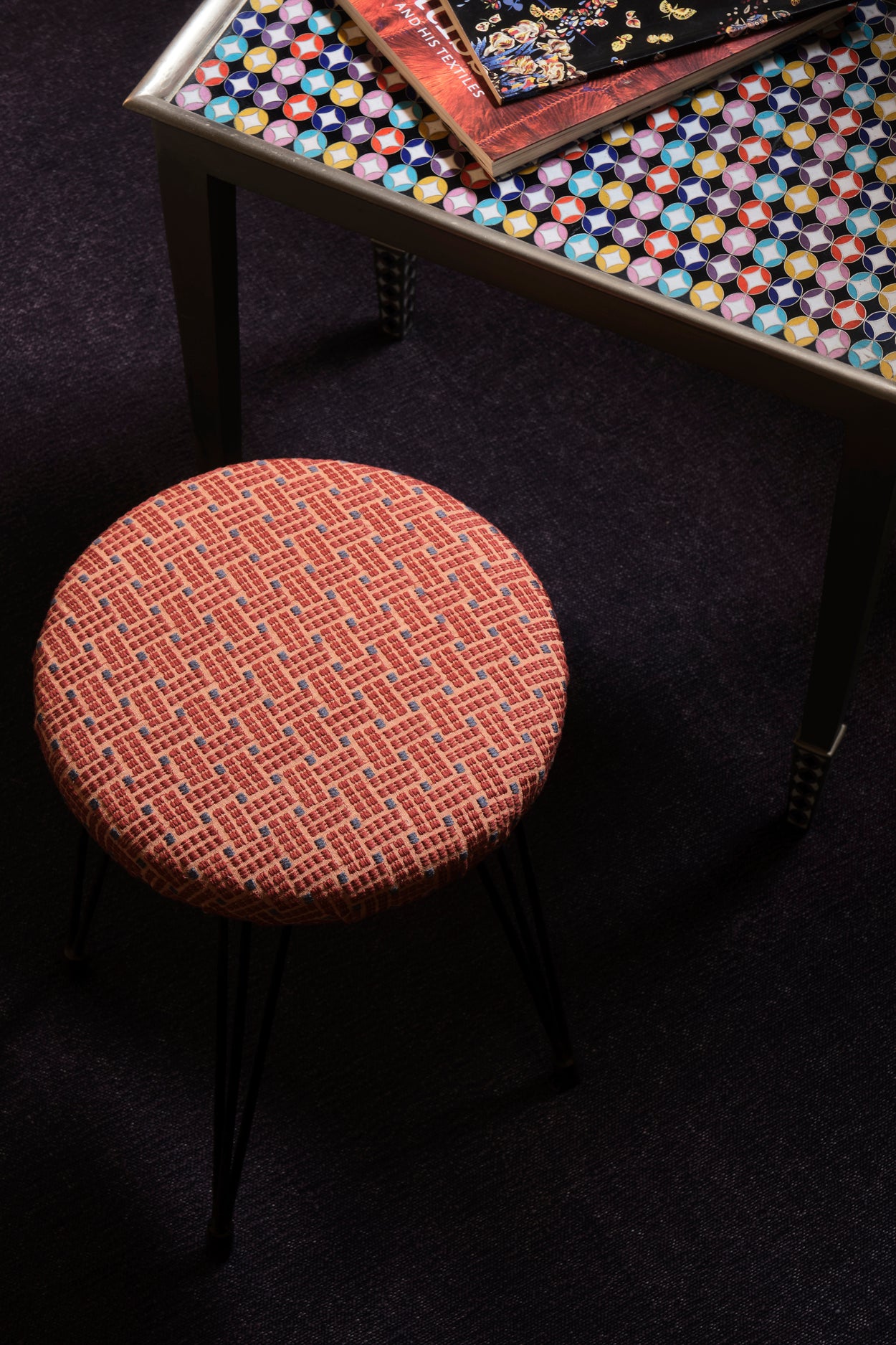 Overhead image of an upholstered stool in a red embroidered fabric with a harlequin enameled side table and stack of books. 
