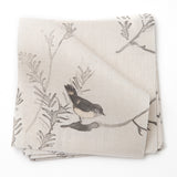 A stack of fabric swatches in a painterly bird and branch pattern in shades of gray on a greige field.