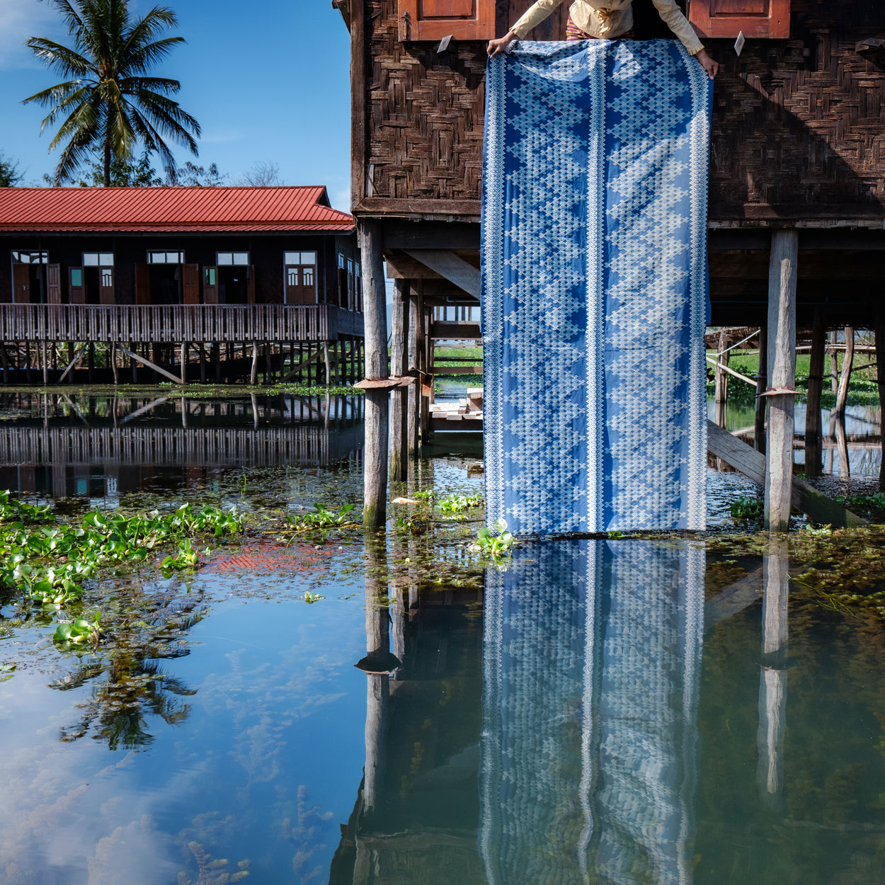 Woman holding a large blue and white patterned fabric in front of a wooden house on stilts on a waterway.