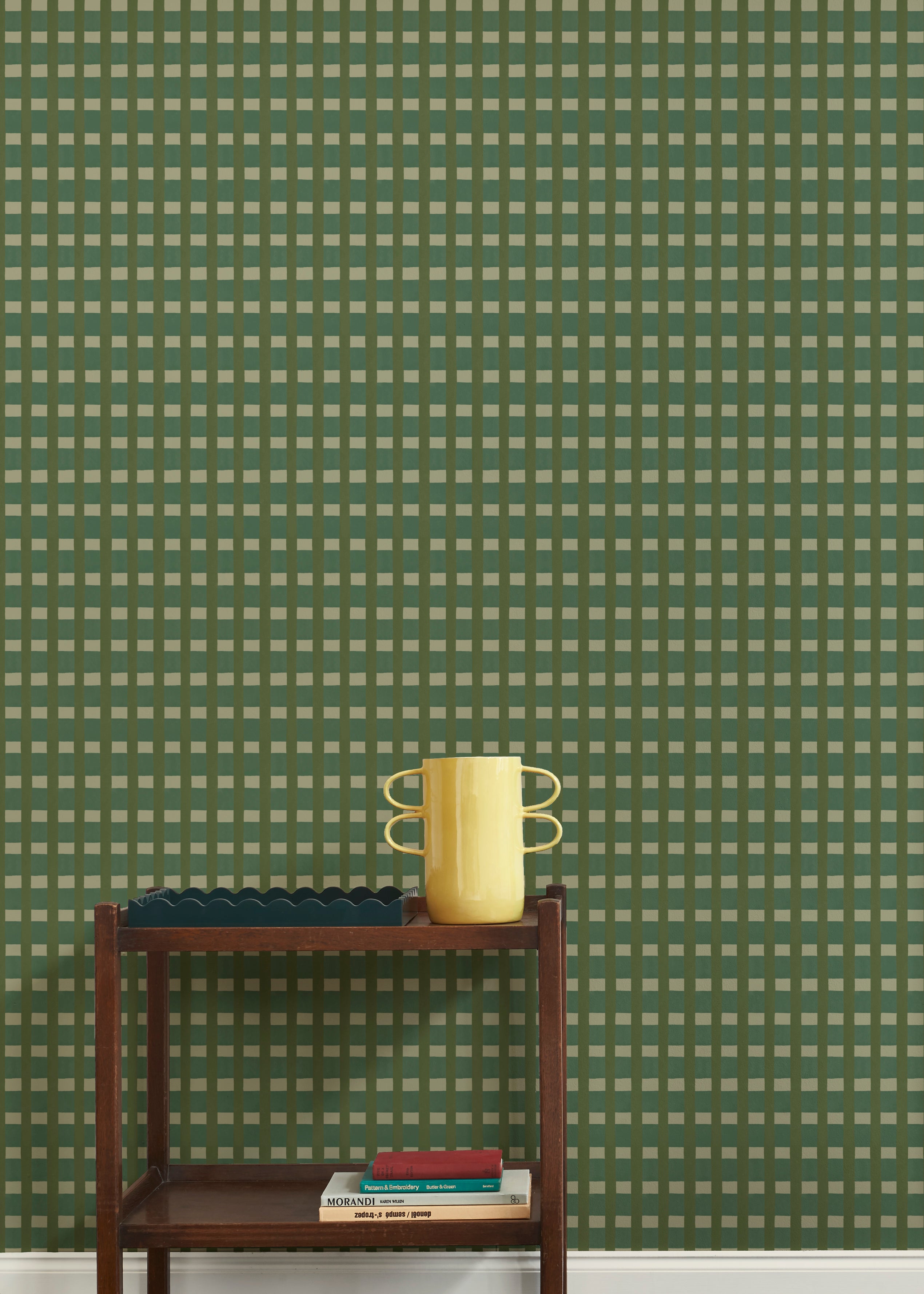 A wallpapered wall in a gridded check pattern in greens with a wooden cart with a yellow vase, green scalloped tray and a stack of 3 books. 