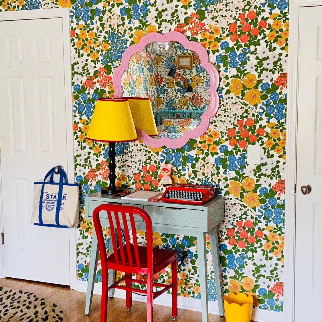 A colorful room corner featuring a retro floral wallpaper wall, a light blue desk, a red chair, a yellow lamp, and a pink scalloped mirror.