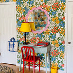 A colorful room corner featuring a retro floral wallpaper wall, a light blue desk, a red chair, a yellow lamp, and a pink scalloped mirror.