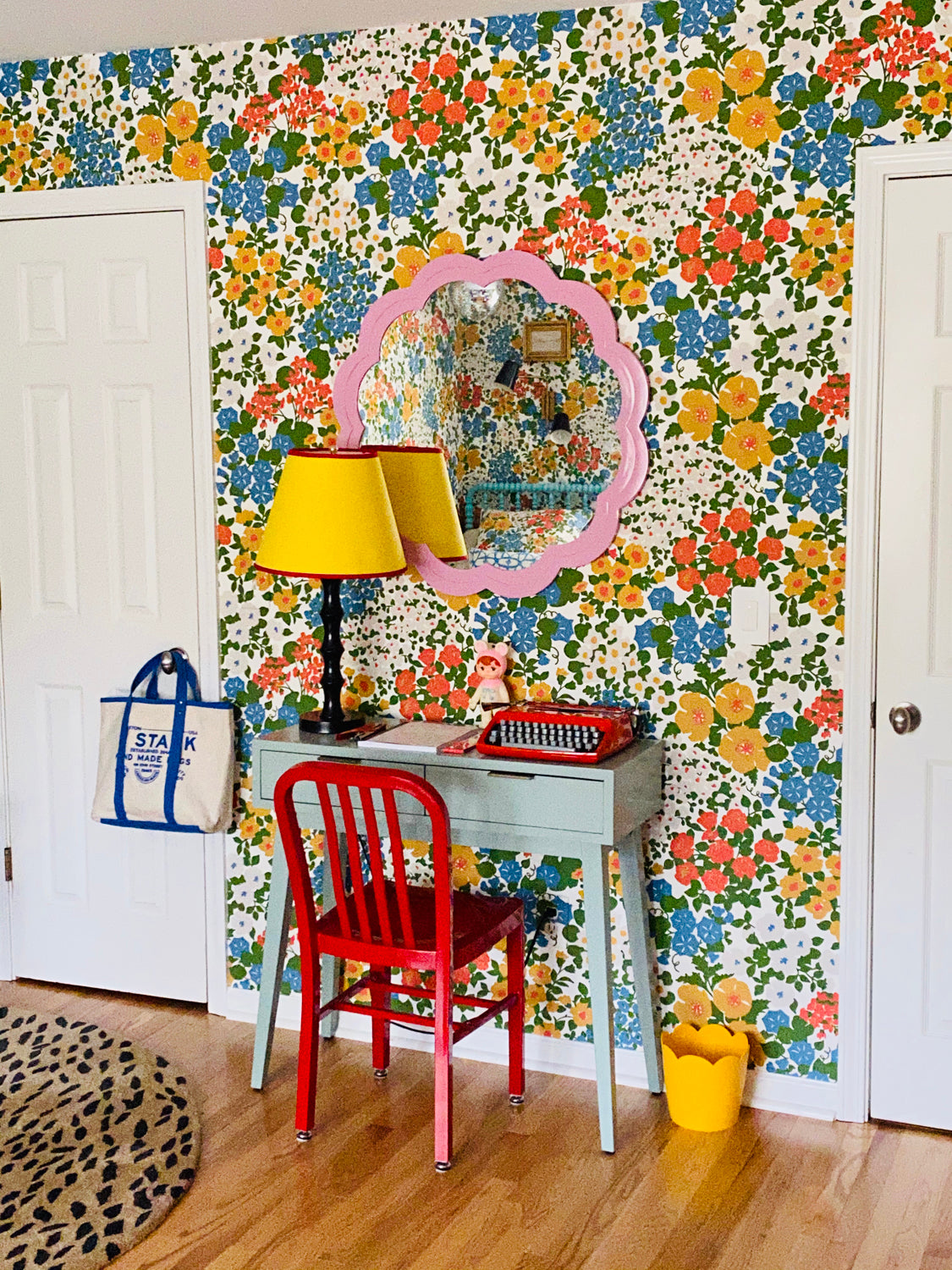 A colorful room corner featuring a retro floral wallpaper wall, a light blue desk, a red chair, a yellow lamp, and a pink scalloped mirror.